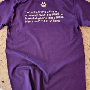 Puede incluir: Camiseta morada con una huella de pata blanca y una cita sobre los animales: "Cuando miro a los ojos de un animal, no veo un animal. Veo un ser vivo. Veo un amigo. Siento un alma." ~ A.D. Williams