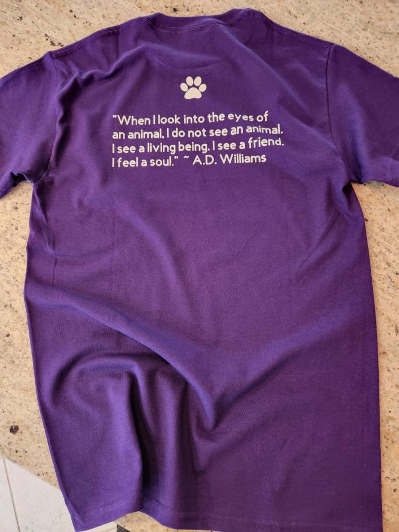 Puede incluir: Camiseta morada con una huella de pata blanca y una cita sobre los animales: "Cuando miro a los ojos de un animal, no veo un animal. Veo un ser vivo. Veo un amigo. Siento un alma." ~ A.D. Williams