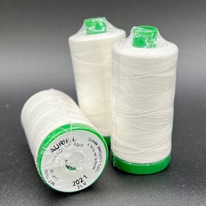 May include: Three spools of white thread with green tops. The thread is labeled "Aurifil Mako NE 40/2 100% cotone Mako Made in Italy 2LV 2021 GR 30 MI 1000"