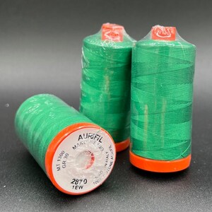 May include: Three spools of green thread with orange and white labels. The labels read "Aurifil Mako 50% Cotton 100% Poly 1EW 2870 MT 1300 GR 30".