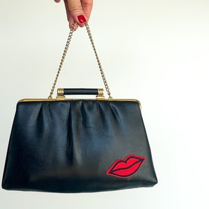 May include: Black handbag with a gold-tone frame and chain strap. The bag features a red embroidered lip design on the front. The handle is black and the bag is held up by a hand.