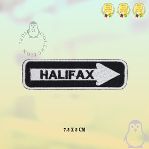 May include: A rectangular embroidered patch with a black background and a white border. The patch features the word HALIFAX in black capital letters centred on a white horizontal arrow pointing to the right. The dimensions are 7.5 x 3 cm.