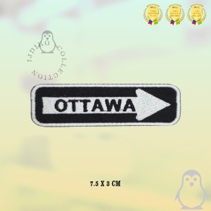 Puede incluir: Parche bordado en blanco y negro con la palabra "OTTAWA" y una flecha blanca apuntando hacia la derecha. El parche mide 7,5 cm por 3 cm.