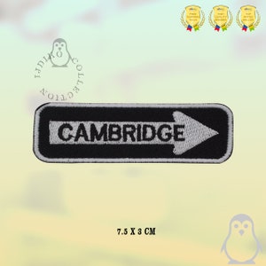 Op de afbeelding: Zwart en wit geborduurde patch met het woord "CAMBRIDGE" en een witte pijl. De patch is 7,5 x 3 cm groot.