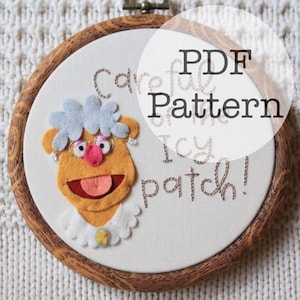 May include: An embroidery hoop containing a felt applique of a yellow character with gray hair, pink glasses, and a white collar. The fabric features embroidered text that reads, Careful of the Icy patch! A circular overlay displays the text PDF Pattern.