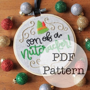 Könnte beinhalten: Gesticktes Stickrahmen-Kunstwerk mit dem Text "son of a Nutcracker!" in schwarzem und grünem Garn. Das Design zeigt einen grünen Elfenhut und goldene Wirbel. Glitzernde Weihnachtskugeln umgeben den Rahmen.