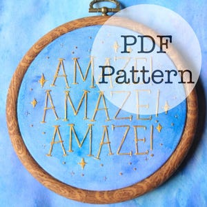 May include: A round embroidery hoop with a blue fabric background. The word "AMAZE!" is embroidered in gold thread. The text "PDF Pattern" is overlaid on the image. The wooden hoop has a brass-colored clasp.
