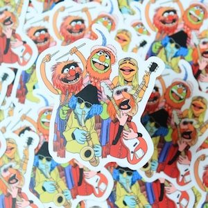 May include: A sticker featuring a group of five Muppet characters playing musical instruments. The characters are Animal, Fozzie Bear, Miss Piggy, Gonzo, and Scooter. Animal is playing the drums, Fozzie Bear is playing the guitar, Miss Piggy is playing the bass, Gonzo is playing the saxophone, and Scooter is playing the keyboard.