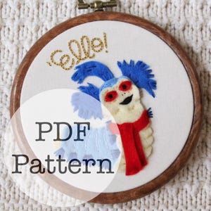 May include: A circular wooden embroidery hoop containing a white fabric panel with a felt applique of a blue and cream-colored creature wearing a red scarf. The word "ello!" is embroidered in gold thread above the creature. The text "PDF Pattern" is overlaid.