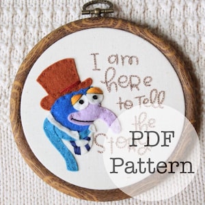 May include: An embroidered hoop art piece featuring a blue character with a long purple nose and a brown top hat. The text "I am here to tell the Story PDF Pattern" is stitched on the white fabric, framed by a wooden hoop.