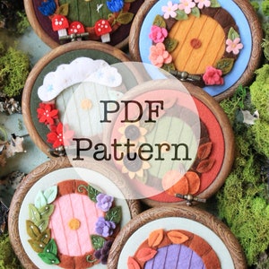 May include: Five felt door wreaths in wooden embroidery hoops. Each wreath features a different color and design, including a green door with blue flowers, a brown door with pink flowers, a white door with a cloud, a green door with sunflowers, and a purple door with fall leaves.