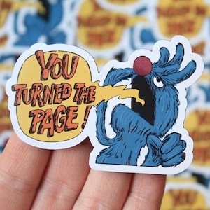 May include: A die-cut sticker featuring a blue, furry, monster-like character with a red nose and an open mouth. A yellow speech bubble extends from the character's mouth containing the orange text, "YOU TURNED THE PAGE!" with a black outline.