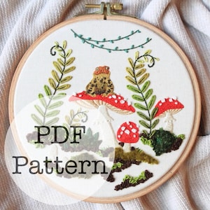 Op de afbeelding: Een PDF-patroon voor een borduurproject met een kikker die op een paddenstoel zit met andere paddenstoelen en groen eromheen. Het patroon zit in een houten borduurring.