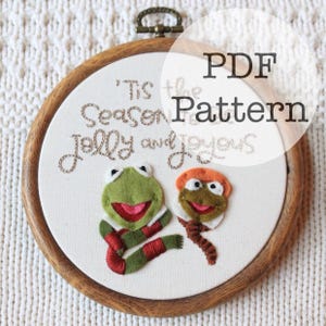 May include: A round embroidery hoop with a cream-colored fabric featuring two felt frog characters and the text "'Tis the Season Jolly and Joyous" and "PDF Pattern". The hoop is made of wood and has a metal hanging loop.