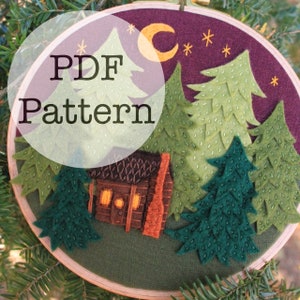 Puede incluir: Un patrón PDF para un proyecto de bordado en fieltro que representa una cabaña en el bosque por la noche. La escena incluye una luna creciente, estrellas y varios pinos. La cabaña tiene un exterior de madera marrón con una chimenea y ventanas. El texto "PDF Pattern" se superpone a la imagen.