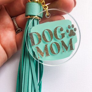 May include: A teal blue acrylic keychain with a gold tassel and the words "DOG MOM" in gold glitter lettering. A gold paw print is also visible on the keychain.