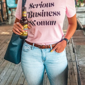 Puede incluir: Una mujer lleva una camiseta rosa con el texto "Serious Business Woman" en letras negras. Lleva pantalones vaqueros azules claros y un cintur&oacute;n marr&oacute;n. Lleva una botella de cerveza en su mano derecha.