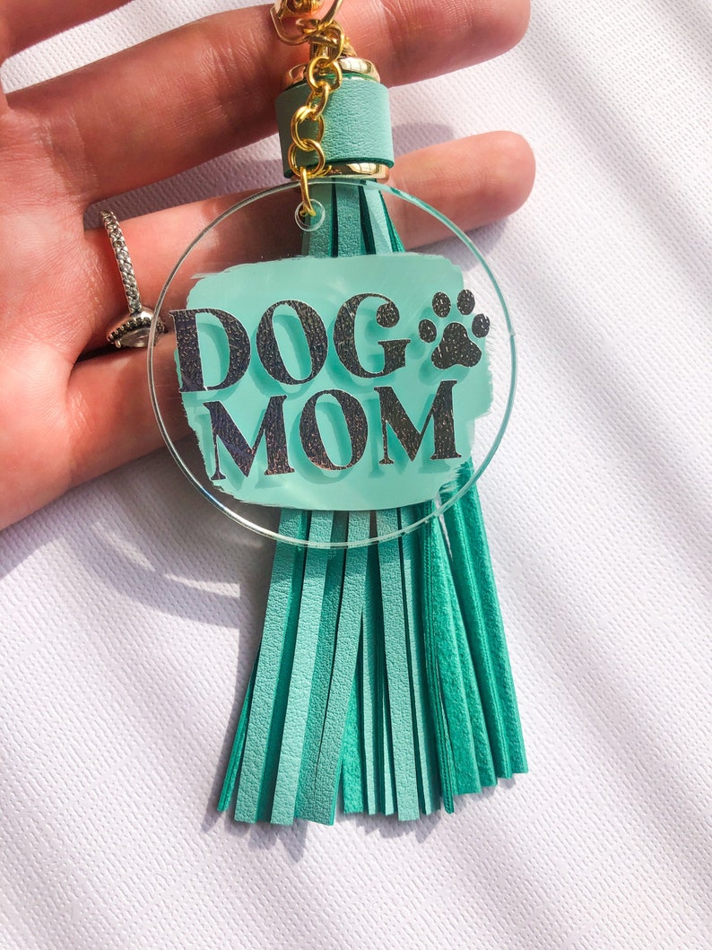 May include: A teal blue acrylic keychain with a silver glitter design that says "DOG MOM" with a paw print. The keychain has a teal blue tassel.