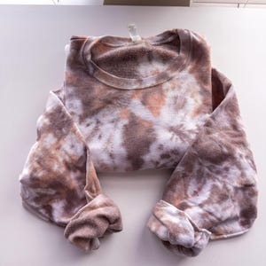 May include: A folded tie-dye sweatshirt in shades of brown, white, and peach. The crew neck sweatshirt has long sleeves with the cuffs folded up. The fabric has a soft, textured appearance.