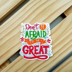 May include: A colorful sticker with the words "Don't be afraid to be great" in a whimsical font. The sticker features red, green, orange, and blue accents. The sticker is placed on a light brown wooden surface.