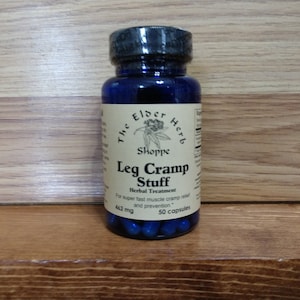 Peut inclure: Une bouteille en verre bleu foncé étiquetée "The Elder Herb Shoppe" contenant "Leg Cramp Stuff", un traitement à base de plantes. L'étiquette indique 462 mg et 50 gélules. La bouteille a un bouchon noir et est sur une surface en bois.