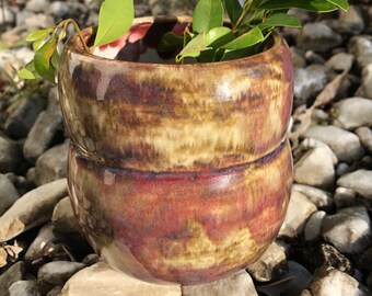 Könnte beinhalten: Eine braune und cremefarbene Keramikvase mit roter Glasur. Die Vase steht auf einem Bett aus kleinen Steinen und hat einen Zweig mit grünen Blättern darin.