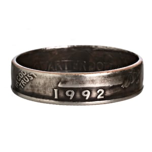 Puede incluir: Un anillo de plata hecho con una moneda de 25 centavos de dólar de 1992. El anillo tiene las palabras "In God We Trust" y "Quarter Dollar" en la banda. El año 1992 es visible en la parte inferior del anillo.