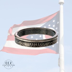 May include: A silver ring made from a vintage Ohio state quarter. The ring features the words "United States of America" and "Ohio 1903" on the band. The ring is displayed against a blurred background of an American flag.
