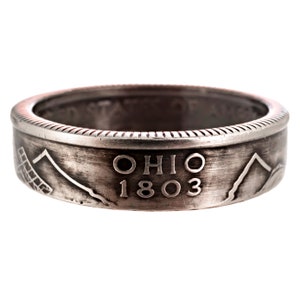 May include: A silver-toned ring made from a repurposed US quarter. The ring features the words "OHIO 1803" and a mountain range design.