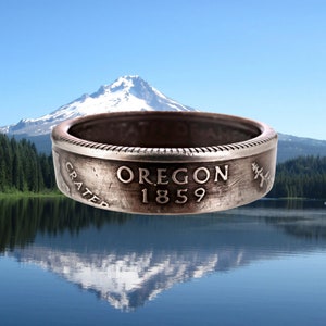 Może przedstawiać: Srebrny pierścionek wykonany z ćwierćdolara z wygrawerowanymi słowami "Oregon 1859" i "Crater". Pierścionek jest wyostrzony na tle rozmytego obrazu góry i jeziora.