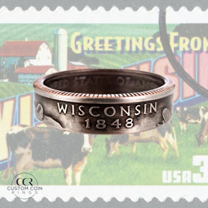 May include: A silver ring made from a half dollar coin with the words "WISCONSIN 1848" engraved on the band. The ring is sitting on top of a vintage postcard with the words "Greetings from Wisconsin" printed on it.