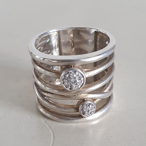 May include: A silver ring with a multi-band design. The ring features two circular accents, each encrusted with small, clear stones. The ring's bands have a textured appearance, creating a layered effect. The ring is a piece of jewelry.