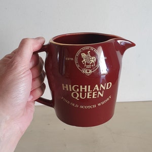 Op de afbeelding: Een rode keramische kan met een gouden rand en de woorden "Highland Queen Fine Old Scotch Whisky" gedrukt op de voorkant. De kan heeft een gouden zegel met een ridder op een paard en de woorden "ESTD" en "QUEEN OF SCOTS" erop gedrukt.