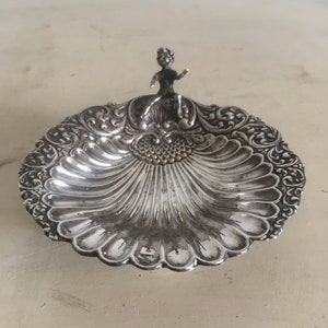 Könnte beinhalten: Eine silberne schalenförmige Schale mit einem verzierten Design und einer kleinen Figur einer Frau, die oben sitzt.