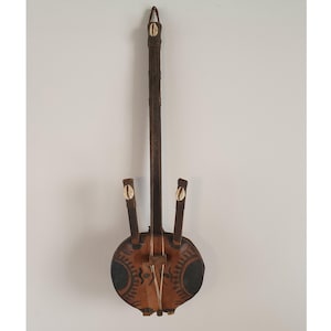 Op de afbeelding: Een handgemaakt snaarinstrument met een donkere houten hals en een ronde, met leer bedekte body. De body heeft decoratieve patronen en twee donkere cirkels. Schelpen sieren het instrument.