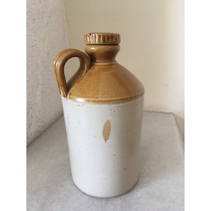 Può includere: Una brocca in ceramica bianca con la parte superiore smaltata di marrone e un coperchio marrone. La brocca ha un manico ed è alta circa 20 cm.