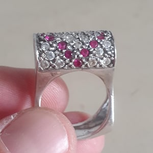 Op de afbeelding: Een zilveren ring met een rechthoekige band bezet met kleine witte en rode edelstenen.