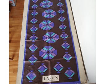 May include: A rectangular brown scarf with a geometric pattern in shades of purple and teal. The scarf has a blue border and the brand name "LANVIN PARIS" is printed in white. The scarf is displayed on a wooden surface.