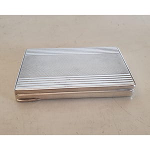 Puede incluir: Un estuche rectangular plateado con una parte superior texturizada y líneas horizontales. El estuche tiene un acabado pulido y un diseño con bisagras. La parte superior presenta un patrón entrecruzado en el centro, rodeado de líneas paralelas. El estuche probablemente se utiliza para guardar objetos pequeños.