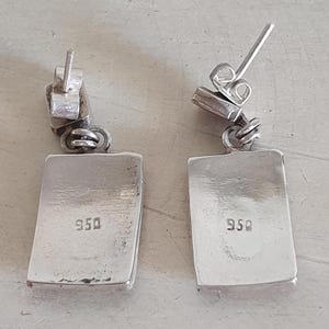May include: A pair of silver earrings with a rectangular shape and a 950 stamp on the back.