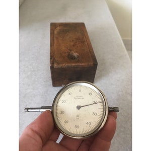 Puede incluir: Un indicador de cuadrante vintage con una carcasa de metal plateado y una esfera blanca. La esfera tiene números y marcas negras, con una aguja que indica una medida. Una caja de madera está en el fondo.