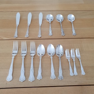 May include: A collection of silver-colored flatware, including butter knives, spoons, and forks, arranged on a wooden surface. The utensils have decorative handles and are likely for formal dining.