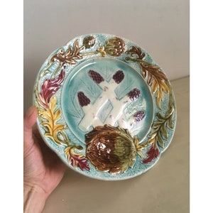 Könnte beinhalten: Ein antiker Teller aus Keramik mit blauer und grüner Glasur. Der Teller zeigt ein erhabenes Design aus Artischocken und Spargel in Braun-, Grün- und Violetttönen.