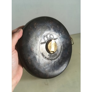 Może przedstawiać: Wstążka miernicza z czarnego metalu w stylu vintage z mechanizmem nawijania w kolorze złotym. Na wstążce mierniczej widnieje napis "Trade Mark" i "Made in the People's Republic of China".