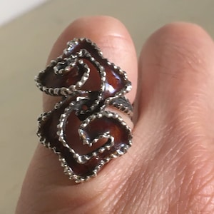 May include: A silver-toned ring with a swirling, floral design. The ring features a dark reddish-brown enamel filling and a textured, beaded edge. The ring is worn on a finger, showcasing its intricate details and craftsmanship.