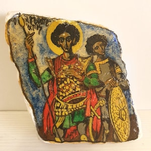 May include: A hand-painted religious icon on a stone. The artwork depicts two figures with halos, one holding a staff with a flame. The figures are dressed in red, green, and gold attire. The background is blue and the stone has a rough, irregular shape.