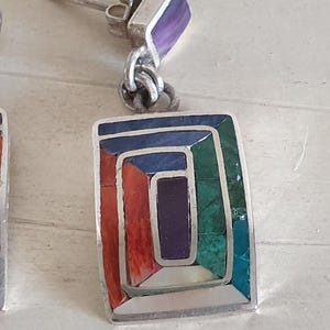 May include: A pair of silver earrings with a geometric design. The earrings are made with a variety of colorful stones, including turquoise, red coral, and purple amethyst.