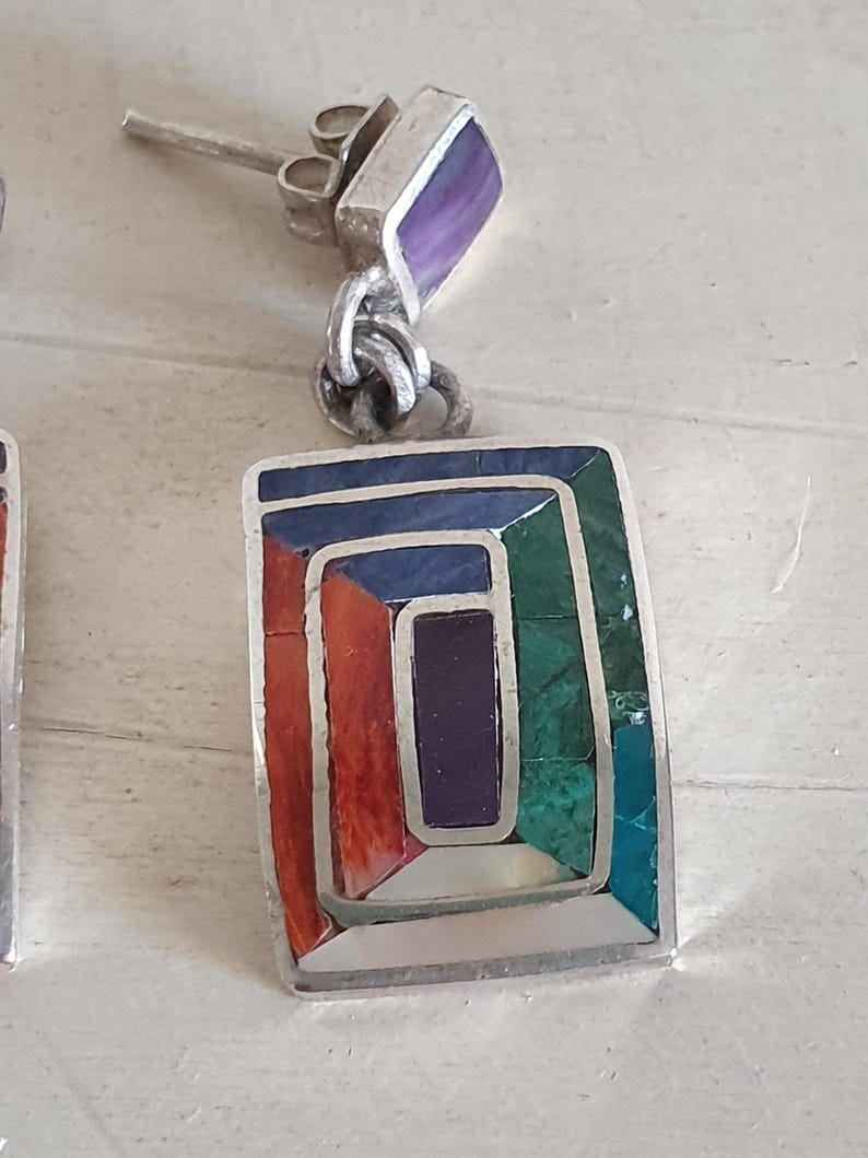 May include: A pair of silver earrings with a geometric design. The earrings are made with a variety of colorful stones, including turquoise, red coral, and purple amethyst.