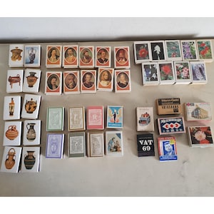 May include: A collection of vintage matchboxes featuring a variety of designs, including images of people, buildings, and abstract patterns. Some of the matchboxes have text on them, such as "VAT 69" and "Wenn Geld..."