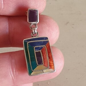 May include: A silver dangle earring with a rectangular shaped stone set in a silver frame. The stone is made of multiple colors of inlaid stones, including green, blue, purple, and orange.